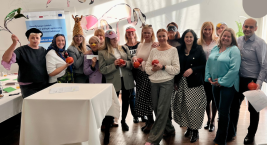 Grupa osób stoi w sali i pozuje z kolorowymi rekwizytami oraz owocami. W tle widoczne są dekoracje ścienne i jasne oświetlenie