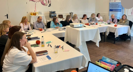 osoby uczestniczące w szkoleniu CUS siedzą przy stołach w sali szkoleniowej, słuchają prowadzącego i robią notatki. Na stołach widoczne są materiały szkoleniowe, owoce oraz rzutnik