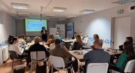 Osoby siedzą przy stołach w sali szkoleniowej i słuchają prowadzącej stojącej przed ekranem z prezentacją dotyczącą współpracy instytucji