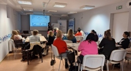 Sala szkoleniowa z kilkunastoma osobami siedzącymi przy stołach, skierowanymi w stronę prowadzącej prezentację wyświetlaną na ekranie projektora.