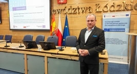 Mężczyzna w eleganckim garniturze w pozycji stojącej przed salą konferencyjną przemawia.