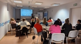 Sala szkoleniowa, w której kilkanaście osób siedzi przy stołach i słucha prowadzącej stojącej przy ekranie z prezentacją.
