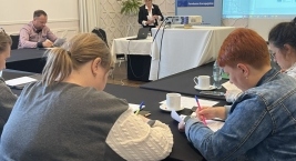 W sali znajduje się grupa osób siedzących przy stołach, skupiona na dokumentach które mają w rękach.