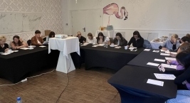 W sali znajduje się grupa osób siedzących przy stołach, zaangażowanych w pracę przy dokumentach.