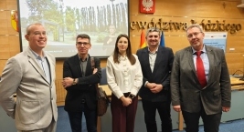 Grupa ludzi stoi przed ekranem, angażując się w prezentację lub dyskusję.  