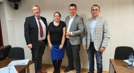 Cztery osoby stoją obok siebie w sali posiedzeń, za nimi ściana, po bokach stoły z notatkami i długopisami